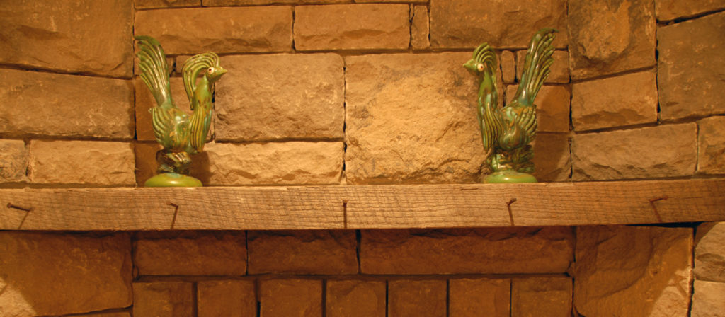 Two charming green ceramic rooster figurines sit on the Rough Sawn 4x12 Fireplace Mantel against a rustic stone wall.
