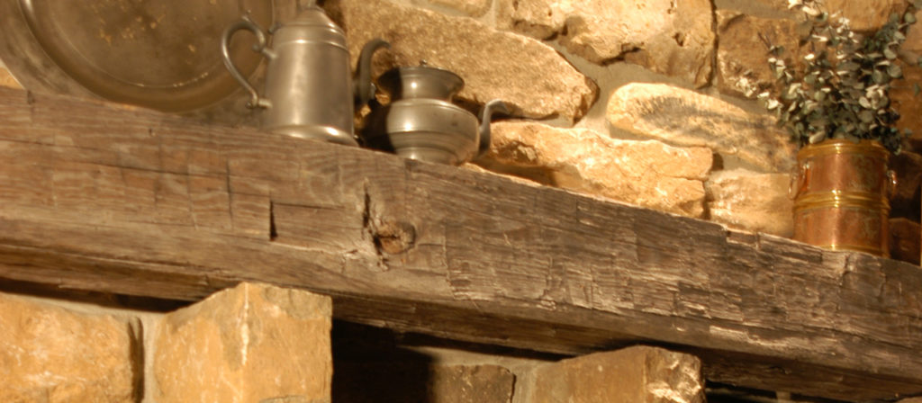 The Hand Hewn Fireplace Mantel 8x8 is a rustic stone mantel featuring a weathered wooden beam adorned with metal teapots and a copper vase with greenery, creating a cozy, vintage ambiance with its warm stone and wood tones.
