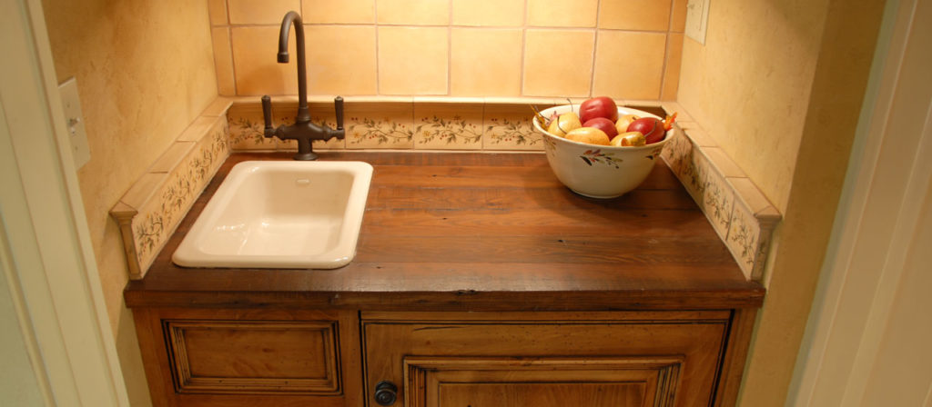 The Rustic Heart Pine countertop, featuring a small white sink and a bowl of assorted apples, pairs beautifully with a floral-patterned tile backsplash, enhancing the warm-toned wall.