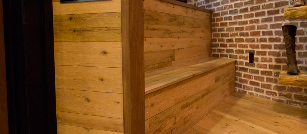 A wooden staircase with a bench, featuring Antique Oak paneling, sits adjacent to a brick wall. A pair of boots on a wooden shelf to the right enhances the warm, rustic ambiance.