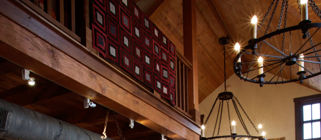 A cozy interior features Reclaimed Antique Rough Sawn Wood Beams and warm lighting, complemented by a geometric pattern quilt draped over the balcony railing. Iron chandeliers with candle-like lights hang from the ceiling, adding a rustic charm to the space.