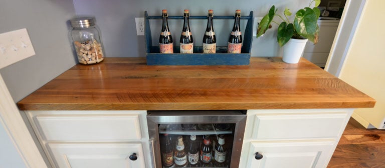 The Coastal Collage home bar setup embraces a coastal vibe with a wooden countertop, featuring five wine bottles in a blue holder, evoking sea colors. A glass jar of wine corks and a potted plant enhance the charm, while a mini-fridge below stores more bottled beverages.