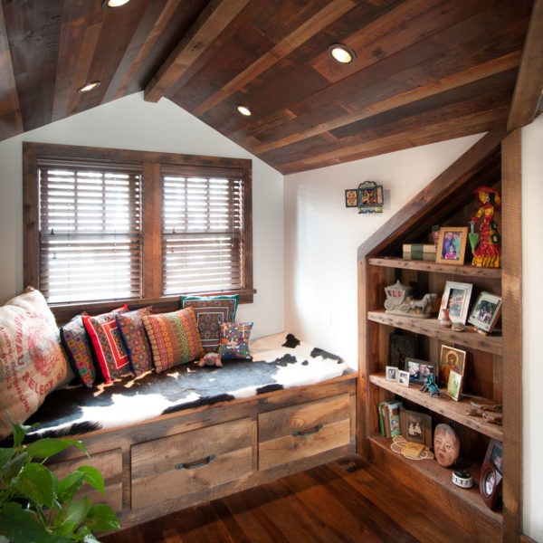 A cozy nook features a wooden daybed with colorful patterned cushions and a cowhide cover under two windows with blinds. A Rough Sawn Shelf 2x8 showcases photos, figurines, and decor. Both the ceiling and floor are wooden.
