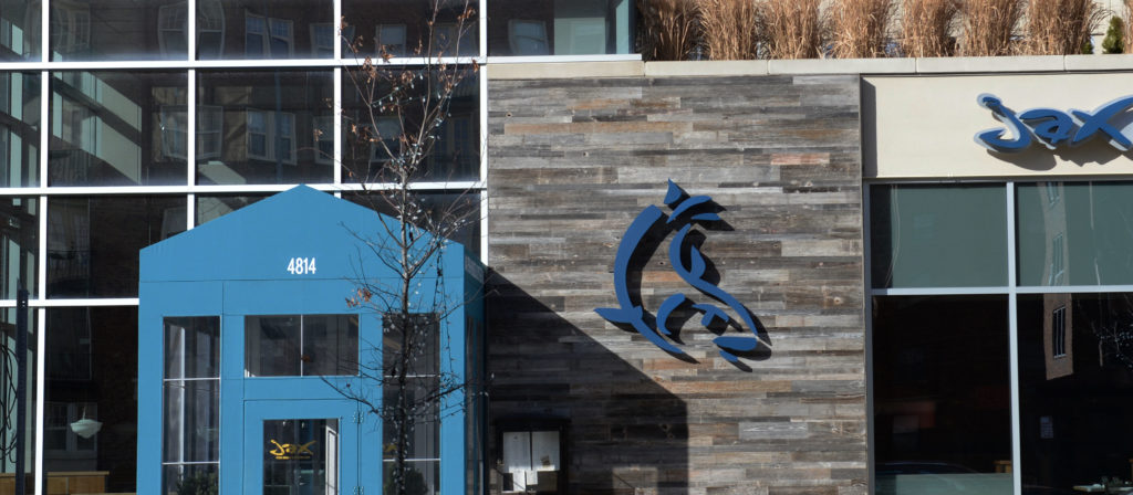 A modern building facade features a blue geometric entrance with the number 4314. Beside it is a textured wall crafted from Reclaimed Grey Barn Wood Siding, complemented by an abstract dark blue metal sculpture. Tall grass elegantly decorates the rooftop edge.