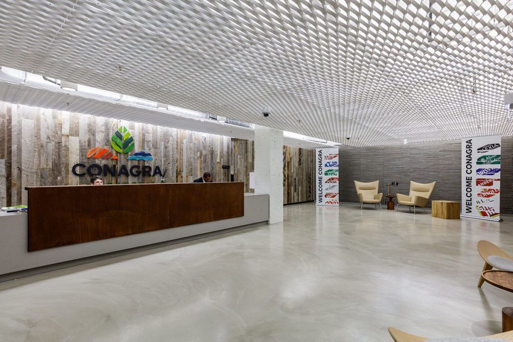 Conagra's reception area boasts a modern look with a wooden desk and accent wall made from Grey Wood Wall Paneling. Beige chairs enhance the neutral palette, while banners feature various logos beneath a ceiling adorned with an eye-catching geometric pattern.