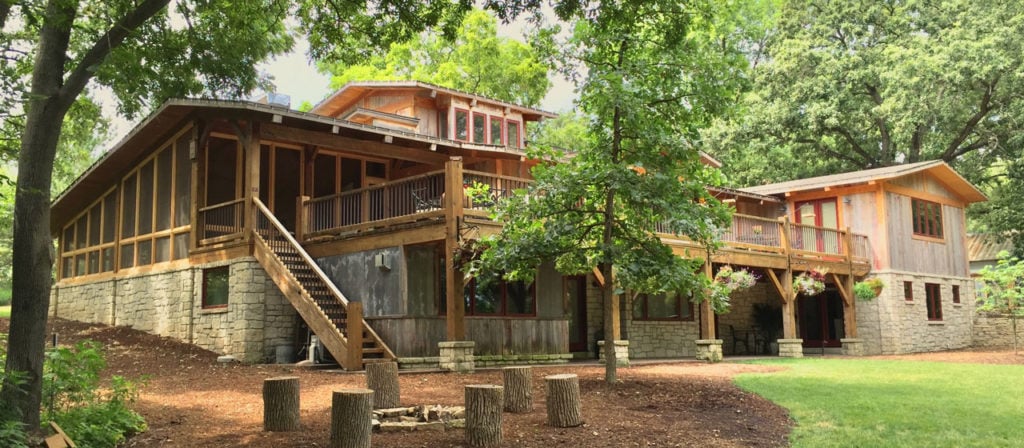 A house with a stone foundation, using Reclaimed Grey Barn Wood Siding, is nestled among trees. It has large windows and a wraparound porch decorated with hanging plants. Tree stumps are artfully arranged on the grassy lawn in the foreground.