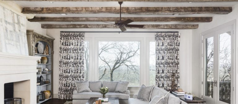 A cozy living room boasts a rustic ceiling with Reclaimed Antique Hand Hewn Wood Beams. Large windows with patterned curtains reveal views of bare trees. The space includes a gray sectional sofa, a fireplace, and decorative shelves.