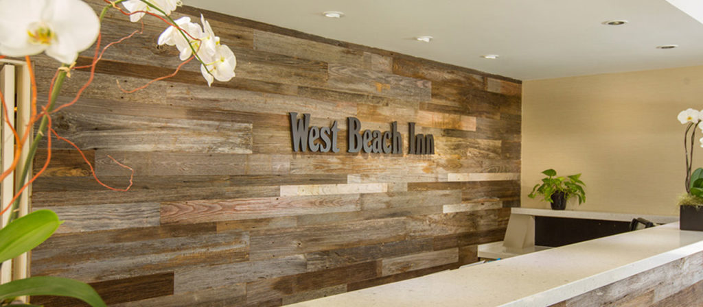 The hotel reception boasts a stunning Grey Wood Wall Paneling with "West Beach Inn" displayed. The reception desk is prominently in the foreground, enhanced by white orchids and lush green plants for a refreshing natural touch.