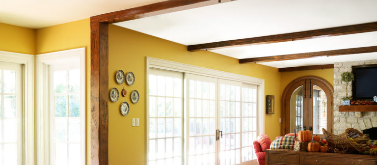 A cozy living room features mustard yellow walls, Reclaimed Wood Box Beams overhead, and a stone fireplace. A round mirror and decorative plates adorn the walls. Sliding glass doors bring in natural light, while a couch with plaid cushions invites comfort.