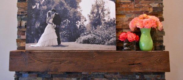 A framed black and white photo of a couple kissing in a garden enhances the rustic charm of the Rough Sawn Fireplace Mantel 6x6. A green vase with pink and orange flowers contrasts beautifully against the stone wall backdrop, adding a vibrant splash of color.