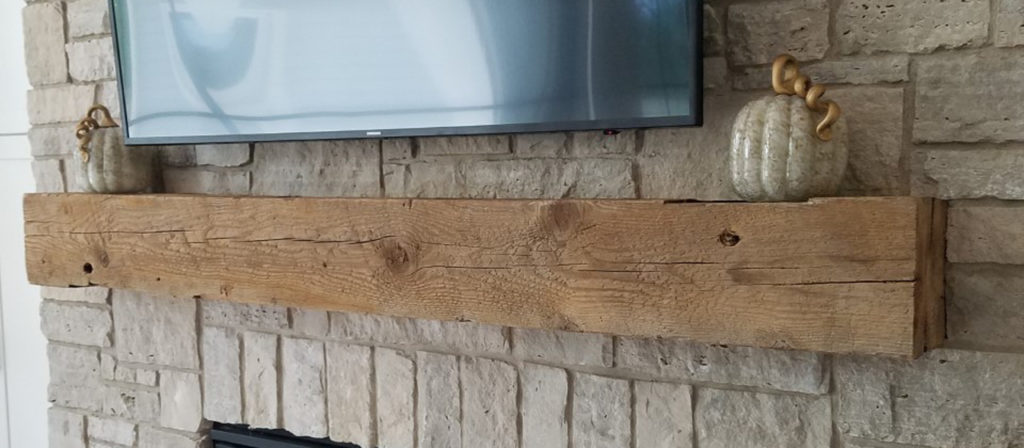 A Rough Sawn Fireplace Mantel 6x6 is mounted on a stone fireplace, with two decorative white pumpkins with curly stems on either side, and a large flat-screen TV positioned above it.