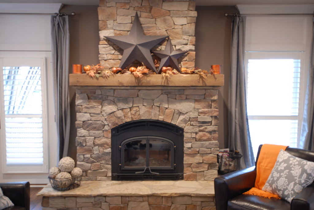 A Rough Sawn Fireplace Mantel 8x8 is adorned with metal stars and autumn garland. It's flanked by tall windows with white shutters. Black leather chairs sit nearby, one draped in an orange blanket, while a basket of pinecones rests on the hearth.