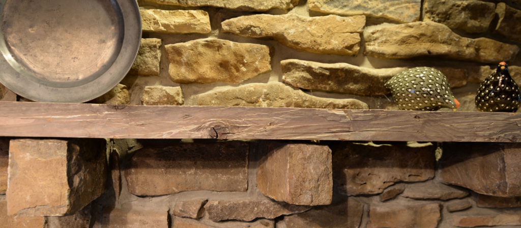 A rustic stone wall features a Rough Sawn Fireplace Mantel 2x8 showcasing a metal plate and two decorative guineafowl figurines, enhancing the cozy, vintage feel.