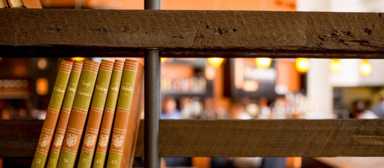 Close-up of Rough Sawn Shelf 2x10 holding brown and green books on the left, set against a softly blurred warm-lit interior, creating an inviting cozy cafe or library atmosphere.