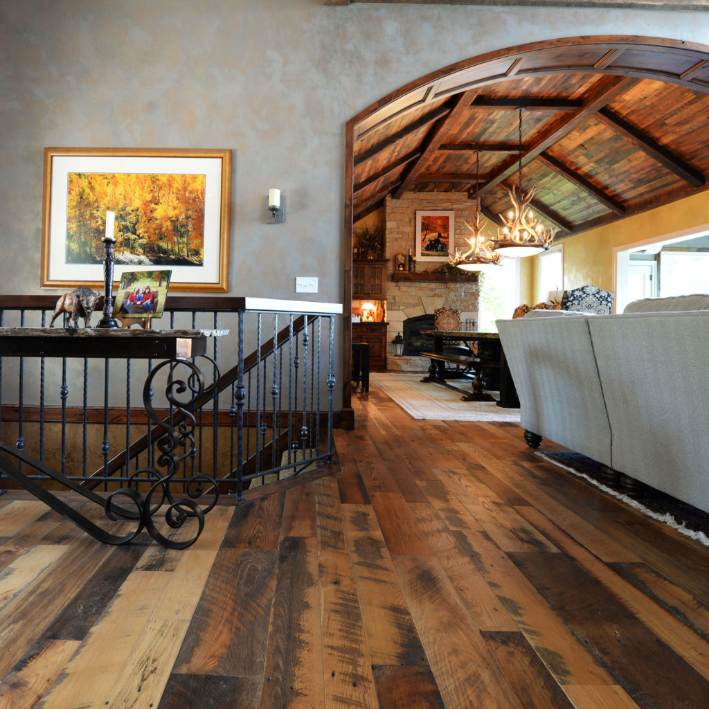 The Coastal Collage rustic living room features a wooden floor, arched wooden ceiling, and stone fireplace for coastal charm. A large sofa faces the fireplace, art adorns the walls, an antler chandelier enhances coziness, and wrought iron railings add texture in the foreground.