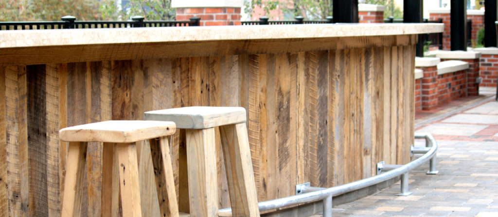 Two Rustic Oak stools sit beside a long wooden bar with a metal footrail, accentuated by rustic oak paneling. The patio area's brick wall and paver flooring complete the cozy outdoor setting.