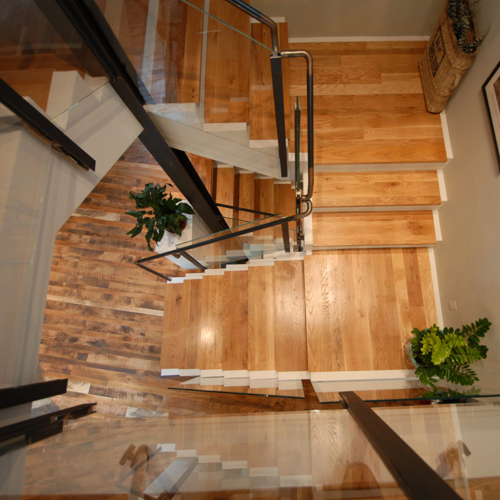 A modern staircase crafted with Matching Stair Treads, Risers and Parts, featuring glass railings and metal supports, viewed from above. Potted plants add greenery on each landing, while lighter wood flooring complements the darker steps beautifully.