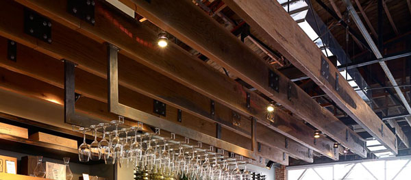 Ceiling-mounted glass rack in a bar or restaurant, featuring wooden beams crafted from reclaimed wood and several hanging wine glasses. The setting boasts industrial-style decor with exposed beams and pipes. Natural light filters through a large window, enhancing the sustainable charm.