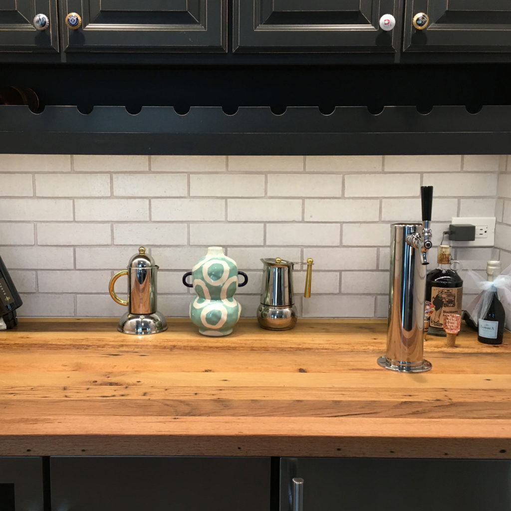 A wooden countertop showcases two metallic coffee makers, a decorative ceramic jug, and a beer tap. Against the white tiled wall and dark cabinets, the design embodies enduring elegance. For those seeking lasting results in wood kitchen countertops, various bottles partially peek into view on the right.