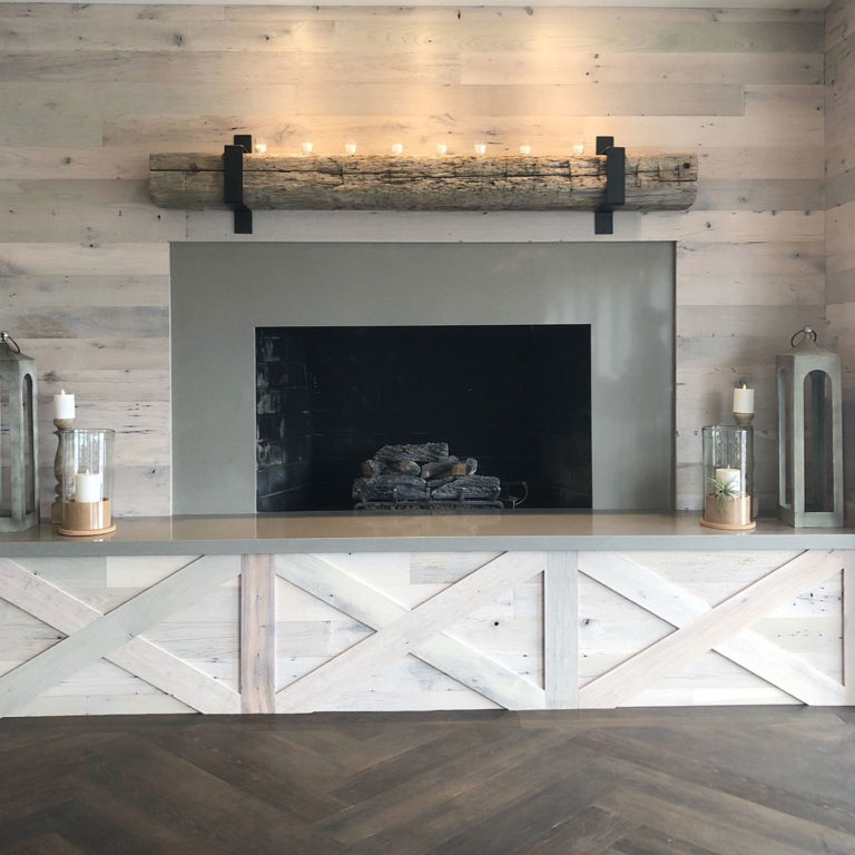 A rustic fireplace set in a wooden wall, topped with a log mantel displaying small candles, shows how to decorate with warmth. Two tall lanterns with candles flank the hearth. The wood paneling has a light, weathered finish, complementing the dark herringbone floor beautifully.