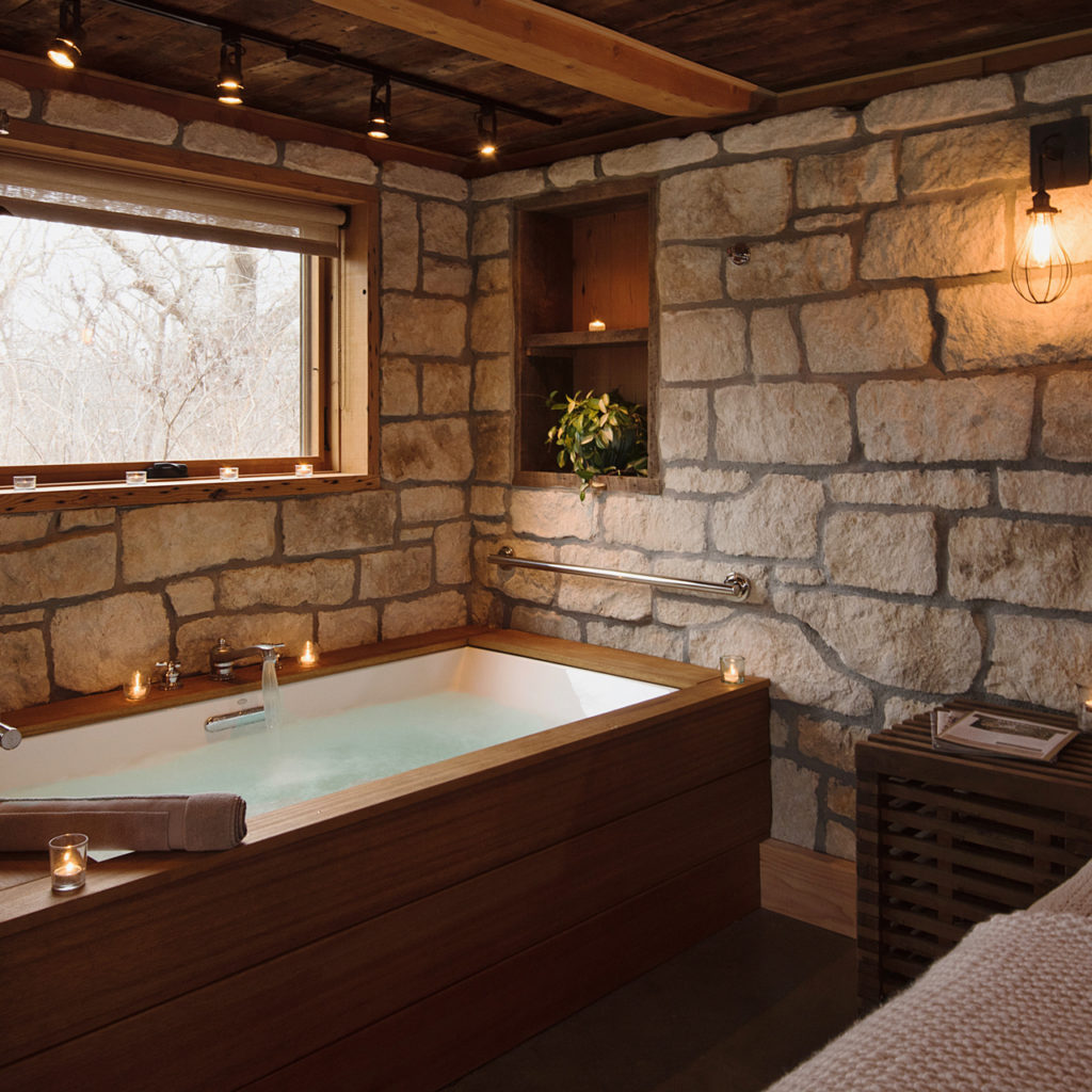 Cozy bathroom featuring stone walls and a wooden soaking tub crafted from reclaimed wood, filled with water. Lit candles are placed around, and a small plant sits in a wall niche. A window offers a view of bare trees, while an exposed bulb provides warm lighting.