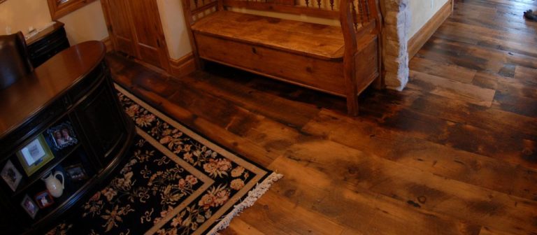A warmly lit room features rustic tobacco wood flooring, a dark wooden desk with framed photos, and an ornate black floral rug. A wooden bench is placed along the wall in front of a stone accent, creating a cozy atmosphere.