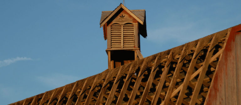 A rustic wooden cupola crafted from reclaimed wood with slatted sides sits atop a weathered roof against a clear blue sky. The roof shows geometric patterns created by wooden beams and slats, showcasing the beauty of recycled materials.