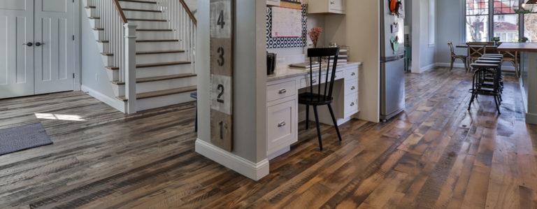 A spacious kitchen and hallway featuring rustic wood flooring, complemented by a white desk with a black chair. The staircase boasts a white railing. Natural light floods the area through large windows near the dining table, highlighting the charm of the dirty top wood flooring.