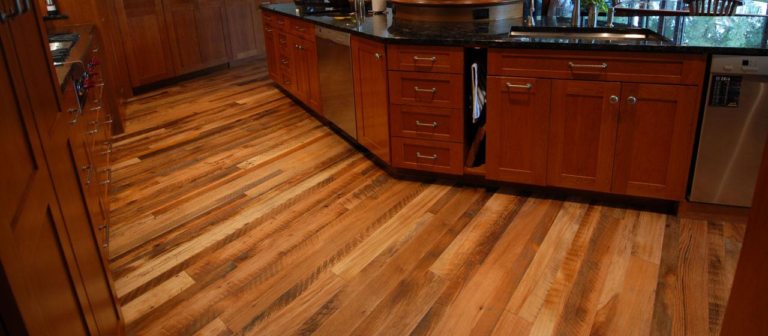 A kitchen with polished wood flooring and matching cabinets exudes warmth. The countertop is black, complementing the stainless steel appliances, showcasing a flawless flooring project.