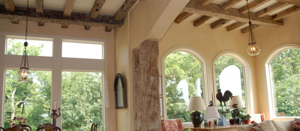 A cozy living room showcases rustic wooden beams crafted from reclaimed wood on the ceiling and large windows overlooking lush green trees. With a sofa, lamps, and decorative items on a table, it offers design ideas for creating a warm, inviting atmosphere.