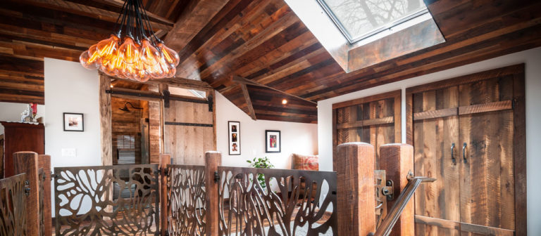 A timeless rustic interior showcases a barnwood ceiling and floor, complemented by a decorative metal railing. The unique chandelier with orange lights, coupled with a skylight, illuminates framed pictures on the walls and wooden double doors. A plant and armchair enhance the cozy ambiance.