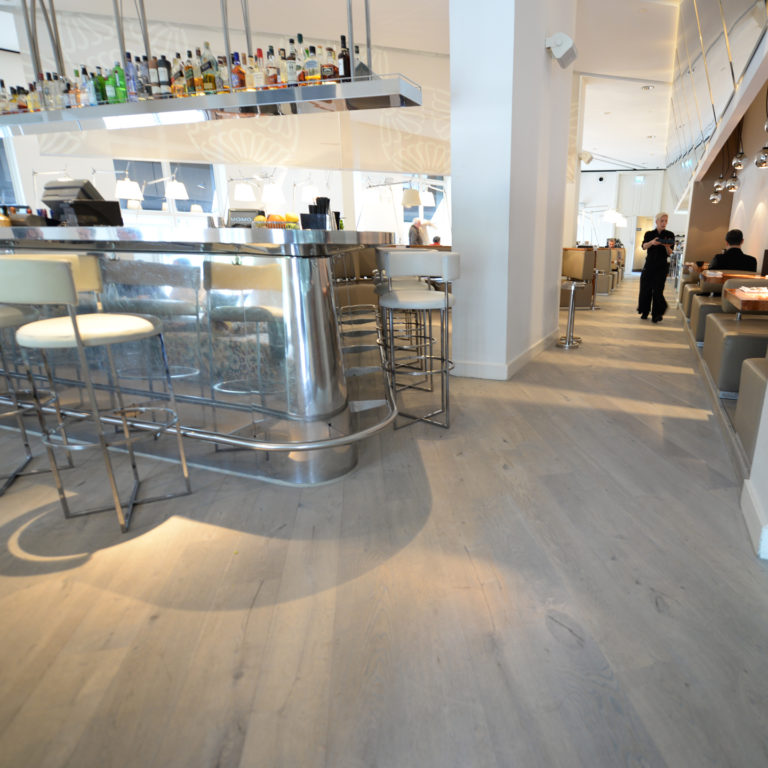 Modern bar interior with a sleek, curved metallic counter and high stools. A range of bottles is displayed above the bar. The room features elegant hardwood flooring and contemporary lighting, with people seated at tables in the background.
