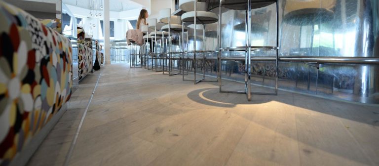 A wide-angle view of a modern, stylish cafe showcases whitewash flooring beneath high metal bar stools and a reflective counter. The left side features a colorful, patterned sofa, while shades of white enhance the ambiance. A person is slightly visible in the background.