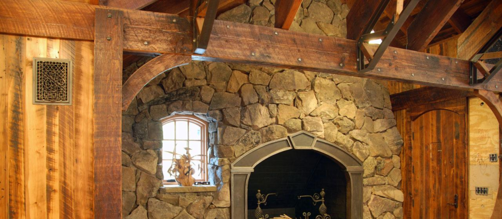 A rustic stone fireplace is set against a wooden wall with exposed beams, evoking charming design ideas. A small, arched window with decorative metalwork sits beside the fireplace, allowing natural light to illuminate the warm, cozy interior enriched by reclaimed wood elements.
