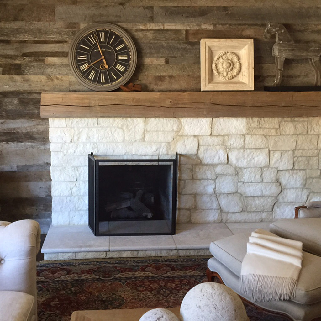 Cozy living room with a rustic stone fireplace and wood wall paneling. A mantel holds a decorative clock, a framed art piece, and a small reindeer sculpture. A patterned rug, two light-colored chairs, and round decorative objects complete the warm setting.
