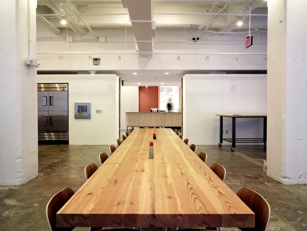 Spacious modern office kitchen with a long wooden communal table surrounded by chairs and a stunning finish. The stainless steel refrigerator on the left complements the countertop in the background, enhanced by industrial-style lighting and beams. Bright, minimalist decor with barn siding details.