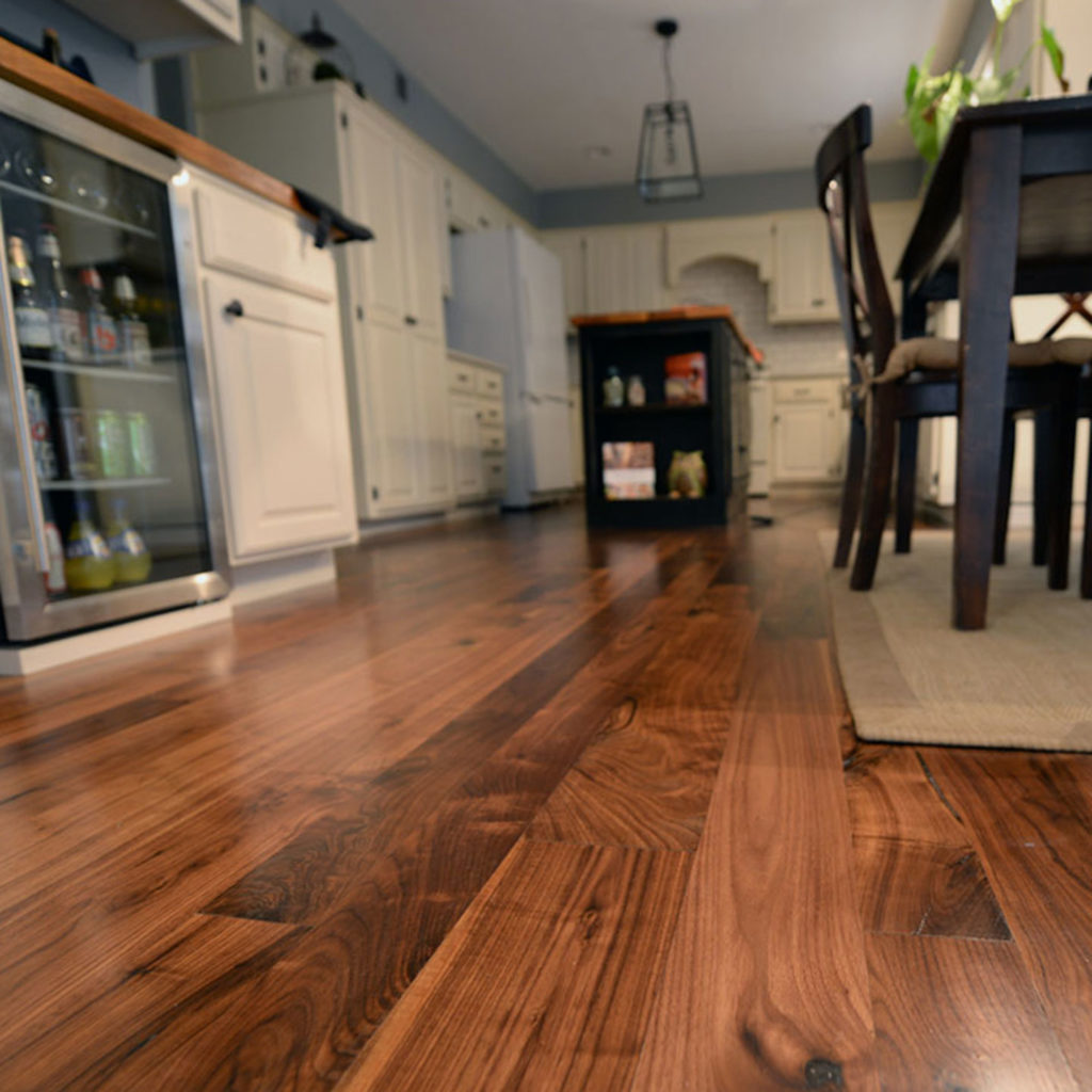 The popular kitchen interior boasts polished walnut hardwood flooring. White cabinets line the walls, and a dining table with chairs is on the right. A glass-door refrigerator is visible, and a hanging pendant light hovers above an island in the background.