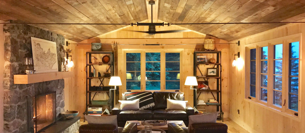 Cozy living room with wooden ceiling and pine paneling on the walls. A lit stone fireplace is on the left, and a large window in the center overlooks trees. A leather couch with white pillows faces forward, flanked by bookshelves and lamps. Warm lighting fills the inviting space.