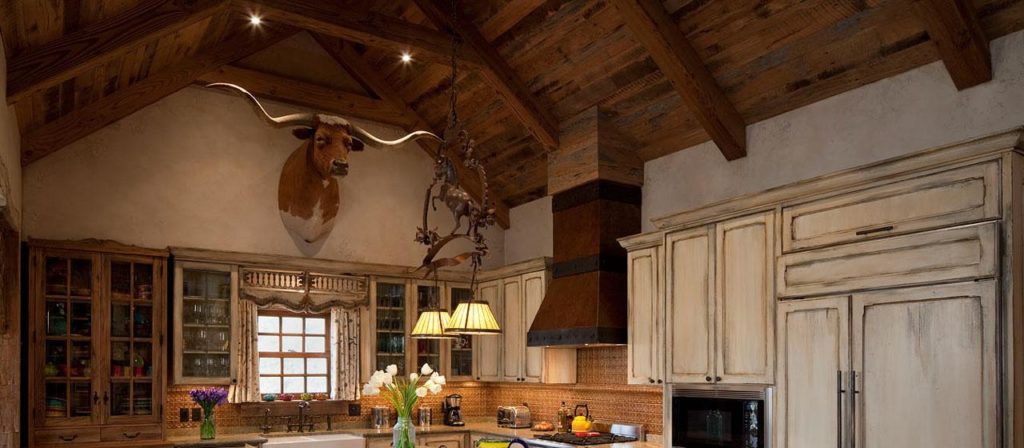 Rustic kitchen with barnwood ceiling beams and distressed cream cabinets. A taxidermy longhorn head decorates the wall above the window. Modern appliances fill the counters, and a timeless chandelier with two lights hangs over the island.
