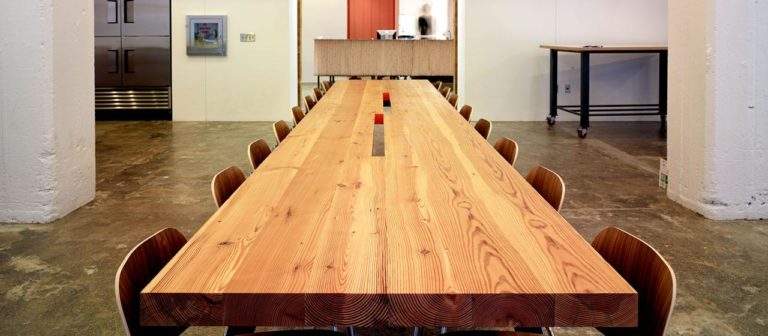 A long conference table crafted from reclaimed wood is surrounded by chairs in a modern office with a concrete floor. A man is blurred in the background near a reception area, while walls and a fridge adorn the left, enhancing the contemporary meeting space ambiance.