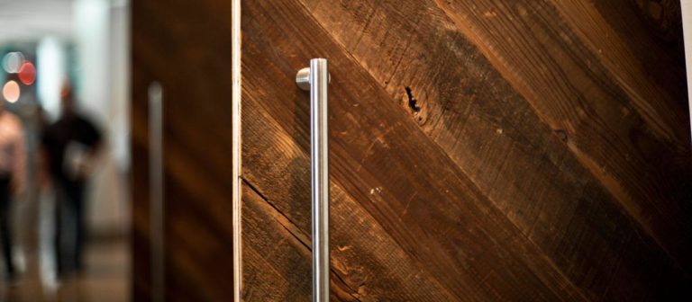 Close-up of a large silver door handle on an angled wall crafted from reclaimed wood, exuding a rustic appearance. The background is blurred, revealing two figures walking in the well-lit hallway of a commercial office interior design.