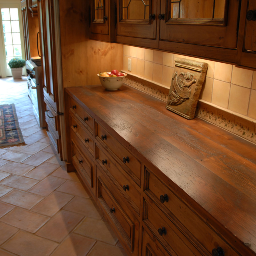 Rustic kitchen with wooden cabinets and wood kitchen countertops. A decorative tile and bowl of fruit sit on the counter. Warm under-cabinet lighting highlights the grain. Tiled floor and a runner rug lead toward a sunlit doorway, showcasing lasting results with a visible plant.