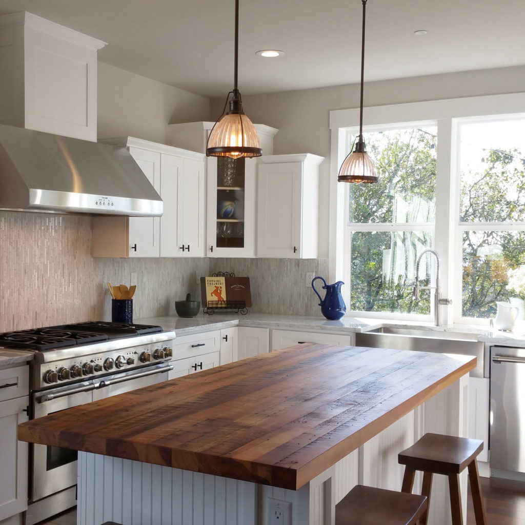 A modern kitchen features white cabinets and a stainless steel stove, complemented by wood kitchen countertops on the large island. Two pendant lights illuminate a blue pitcher near the double sink, framed by a window with views of lush greenery—timeless design ideas for lasting results.