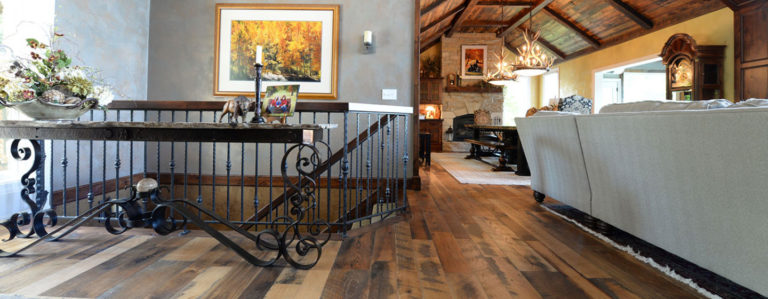 A cozy living room with reclaimed hardwood flooring and a vaulted ceiling. There's a sofa on the right, a grand piano near a stone fireplace, and framed artwork on the walls. A decorative wrought-iron railing leads down a staircase. Natural light fills the space.