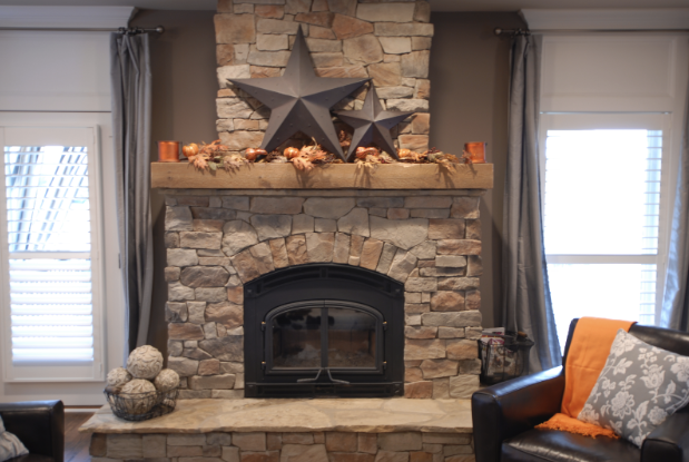 A rustic stone fireplace with a reclaimed wood mantel decorated with two large metal stars and autumn foliage. The hearth has decorative spheres. Dark curtains frame the cozy nook, offering reasons to snuggle in the armchair draped with an orange blanket on a chilly Houston evening.