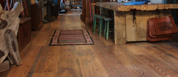 A rustic interior features reclaimed wood flooring, a colorful striped rug, a wooden table with a natural edge, green and wooden stools, and paper bags hanging on the table. The room has a cozy, vintage atmosphere with an inviting weathered look.