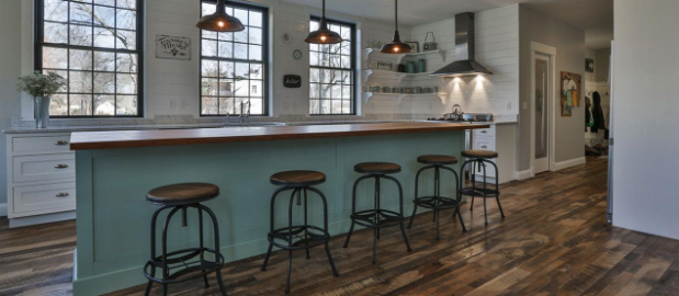 This kitchen showcases a spacious island with a wooden countertop and five industrial-style stools. Under the warm glow of pendant lights, it features two large windows. The wooden flooring adds charm, complemented by a range hood and open shelves in the background, epitomizing modern wood floor ideas.