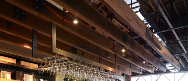 A modern ceiling with exposed wooden beams crafted from reclaimed building materials and industrial lighting fixtures. Upside-down wine glasses hang from a rack, while books rest on a shelf. Skylights, designed by architects in 2019, allow natural light to stream in effortlessly.
