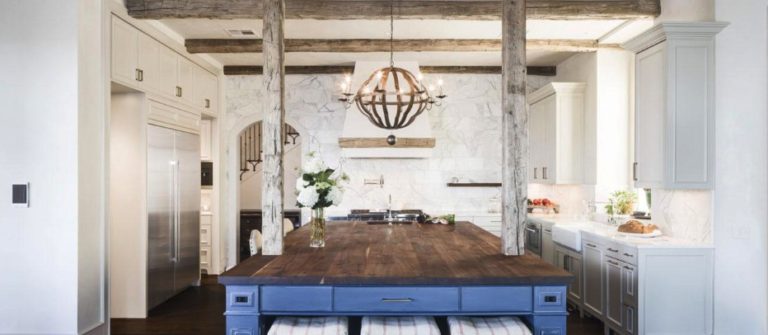 A modern kitchen with a rustic look, featuring exposed wood beams and a large wooden island at its heart. The island boasts blue cabinets, while stainless steel appliances and white cabinetry add contrast. A round chandelier completes the inviting atmosphere.