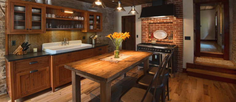 A rustic kitchen features wooden cabinets and a reclaimed wood kitchen table with chairs. A brick backsplash behind the stove adds to the decor, with a vase of orange flowers as a unique focal point. The room is warmly lit by pendant lights, while an entryway opens in the background.